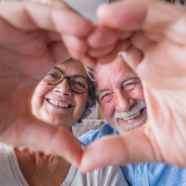 Close up portrait happy sincere middle aged elderly retired family couple making heart gesture with fingers, showing love or demonstrating sincere feelings together indoors, looking at camera.